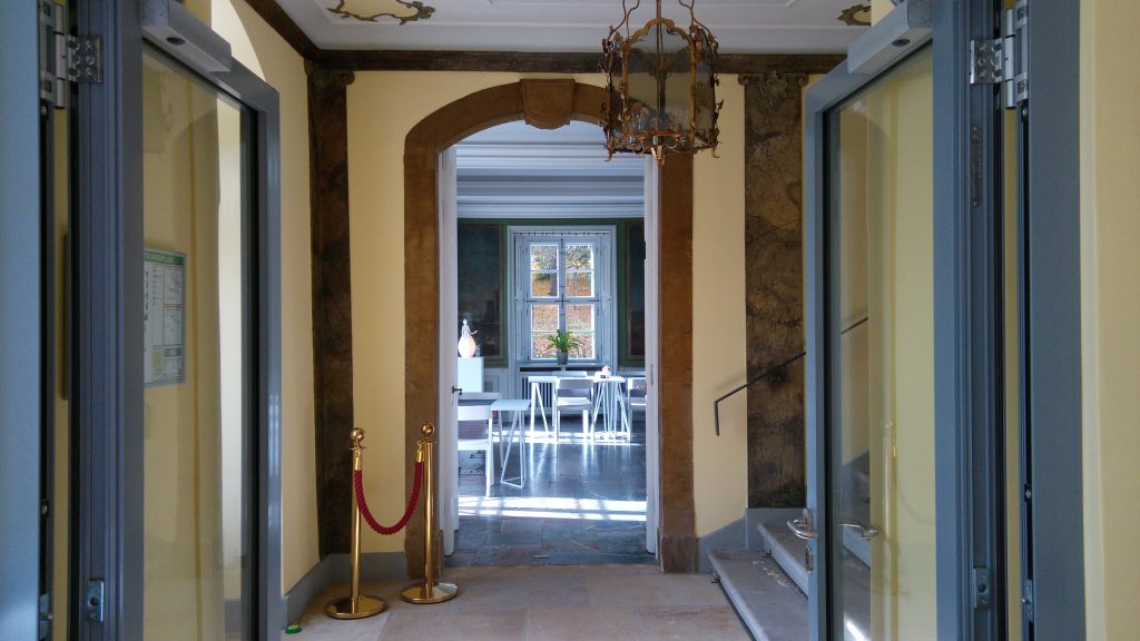 eine offene Tür gibt den Blick in einen angrenzenden, historischen Raum frei, in dem die Sonne scheint und einige Tische stehen. In  der Mitte dort: ein altes Fenster.  