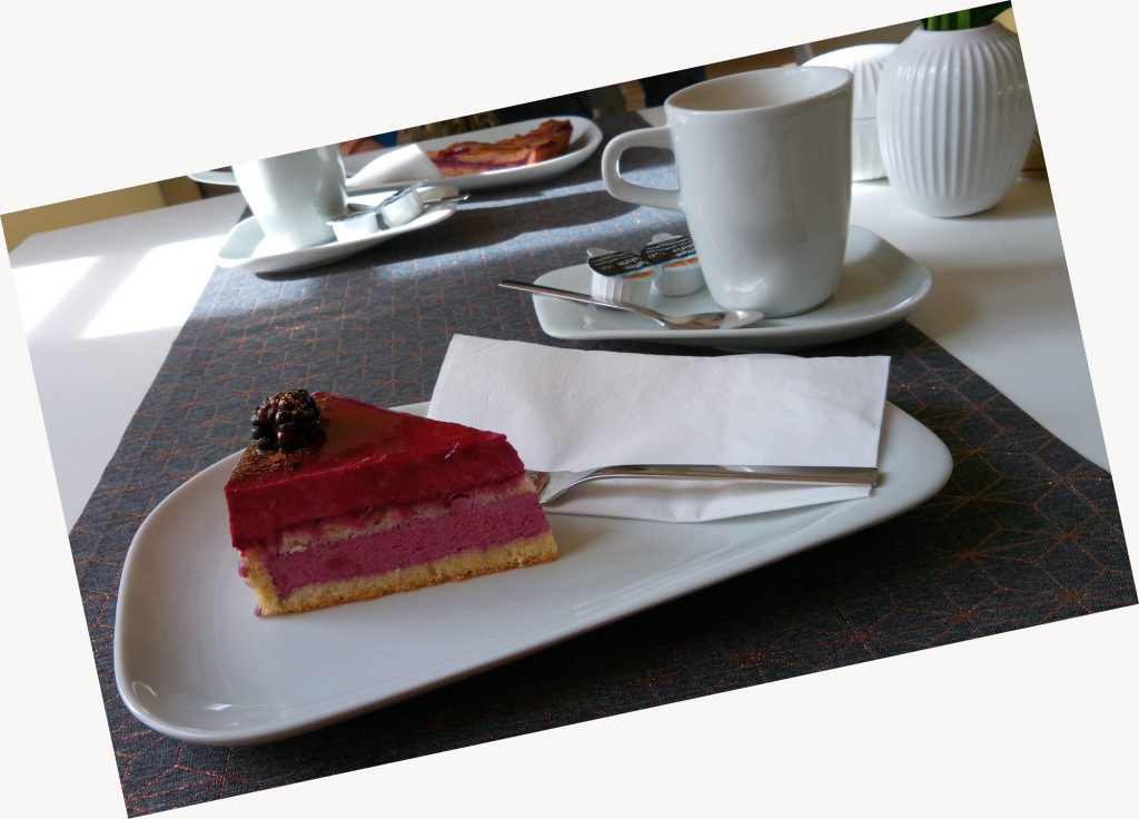 zwei Gedecke mit Torten, dazu ein grauer Tischläufer. Die vordere Torte ist himbeerfarben mit einer großen Brombeere verziert. Dazu Kaffeetassen in weiß. 